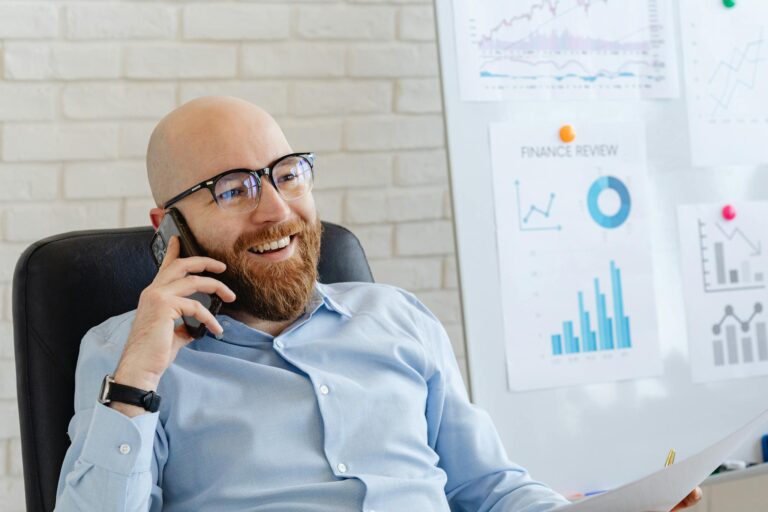 Bald businessman in glasses talks on phone, smiling at office with charts.