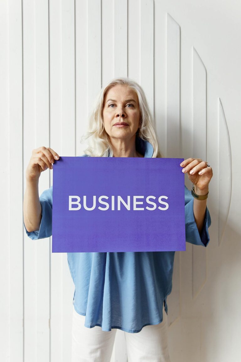 Confident senior woman presenting a business sign in a bright studio setting, wearing a casual blue outfit.
